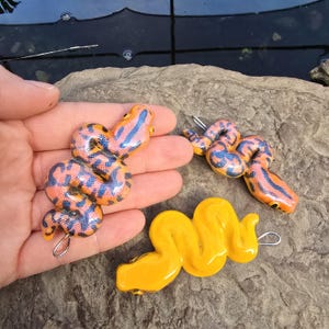 Może przedstawiać: Trzy ręcznie robione wisiorki w kształcie węża. Dwa są pomarańczowo-niebieskie, ze wzorem niebieskich pasków na pomarańczowej podstawie. Trzeci jest w kolorze żółtym. Każdy ma srebrną pętlę do zawieszenia. Wisiorki leżą na szarej kamiennej powierzchni.