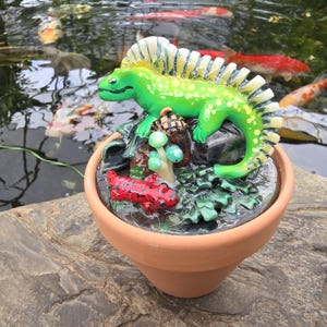 May include: A terracotta pot containing a sculpted green iguana with a spiky crest, a small red salamander with black spots, a brown acorn, and light green mushrooms. The scene is set on a dark, textured base within the clay planter.