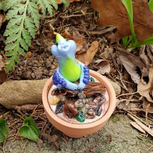May include: A small, blue and green ceramic figurine of a creature sitting on a log in a terracotta pot. The pot is filled with brown and green clay, and there are small blue beads around the creature's neck.