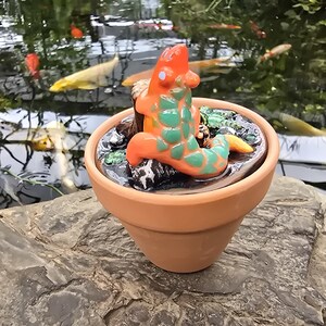 May include: A small terracotta pot containing a glossy, dark, textured surface with a sculpted orange and green lizard-like creature perched on a dark brown log. The creature has a smooth orange body with green scale-like markings on its back.