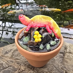 May include: A terracotta pot containing a sculpted scene with a bright pink and yellow salamander resting on dark rocks, brown logs, small yellow spheres, and green moss-like textures. The surface of the scene has a glossy, wet-look finish.