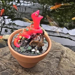 May include: A bright pink lizard figurine with green spots perched on a brown textured log inside a terracotta pot. The base of the pot contains a dark, glossy surface with small green leaf details and a single white flower with a yellow center.