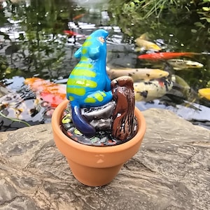 May include: A small terracotta pot containing a sculpted figurine of a bright blue creature with lime green stripes, sitting on a gray rock next to a brown textured object, with a base of small multicolored pebbles inside the pot.