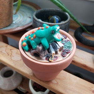 May include: A ceramic incense burner shaped like a pot with a green and yellow lizard on top. The lizard is perched on a pile of logs and leaves. The pot is decorated with a brown glaze.