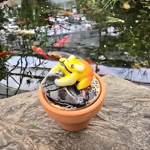 May include: A small terracotta pot containing a decorative figurine of a yellow and orange frog perched on a grey stone. The frog has a smooth, glossy finish and is positioned centrally within the circular opening of the brown clay container.