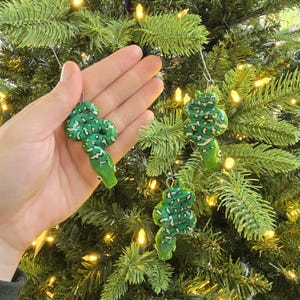 Może przedstawiać: Ręcznie malowane zielono-białe ozdoby choinkowe w kształcie węża. Ozdoby mają wzór tekstury i pętlę do zawieszenia. Są prezentowane na zielonej choince z małymi białymi światełkami.