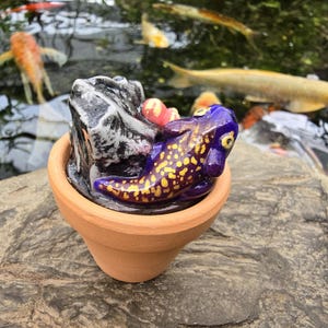 May include: A small terracotta pot containing a sculpted purple salamander with yellow spots resting next to a textured gray rock. The salamander has prominent eyes and a smooth, glossy finish, while the pot has a simple, unglazed clay surface.