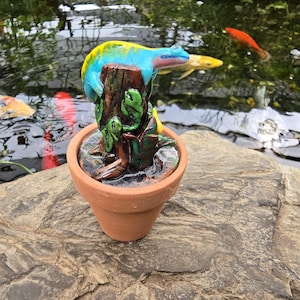 May include: A terracotta pot containing a sculpted brown tree stump with a bright blue and yellow lizard resting on top. The sculpture features green leaves and vines wrapped around the base of the stump, creating a detailed, multi-colored garden ornament.