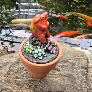 May include: A small terracotta pot containing a sculpted orange creature sitting on a dark stone. The base is decorated with clusters of small, rounded, multi-colored succulent-like shapes in shades of green, light blue, and purple.