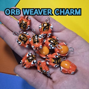 Op de afbeelding: Een verzameling oranje en zwarte spincharms met zilveren springringen. De charms zijn gevormd als spinnen met acht poten en hebben zwarte en witte details. De charms worden op een hand getoond.