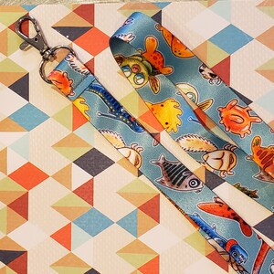 May include: A blue and white lanyard with a repeating pattern of colorful fish and sea creatures. The lanyard has a silver metal clip at the top.