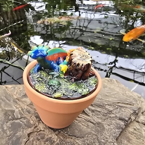 May include: A small terracotta pot containing a miniature scene with a blue and orange lizard-like creature resting on a textured green mossy surface next to a brown tree stump. The surface inside the pot is detailed with small yellow flowers.
