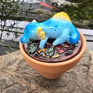 May include: A small blue glass lizard figurine with yellow accents on its head and back, resting on a dark brown circular base with sculpted leaf details, all contained within a simple terracotta-colored clay pot.