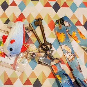 May include: A blue and white plush chicken toy with a red beak and red feet. The toy is attached to a blue and white lanyard with a colorful cartoon design. The lanyard has a silver key chain with two antique keys.