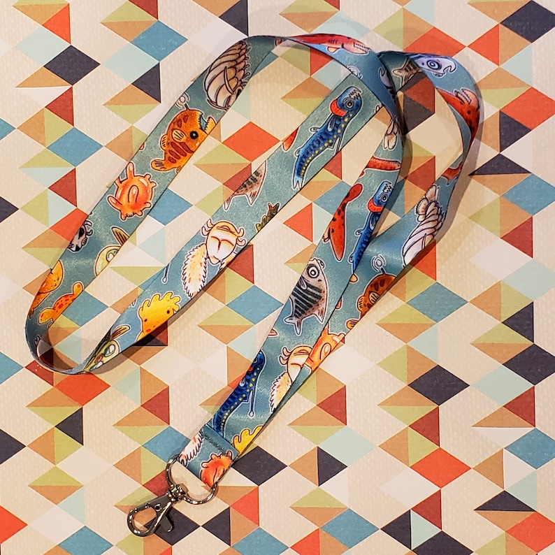 May include: A blue lanyard with a white background featuring a repeating pattern of colorful fish and sea creatures. The lanyard has a silver clip.