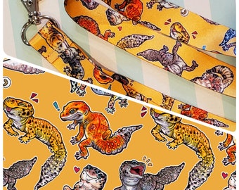 May include: A yellow lanyard with a repeating pattern of colourful geckos. The lanyard is made of a woven fabric and has a black plastic clip.