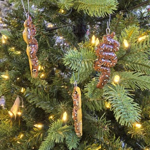 May include: Three handmade Christmas tree ornaments shaped like snakes. The ornaments are brown with white spots and yellow bellies, hanging from a green Christmas tree with white lights.