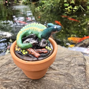 May include: A small terracotta pot containing a sculpted green and yellow lizard resting on dark rocks and brown logs. The lizard has a smooth, glossy finish with a prominent crest along its back, perched atop a circular base inside the clay container.