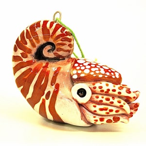 May include: A ceramic nautilus shell ornament with a brown and white striped pattern. The ornament has a green string for hanging and a detailed eye. The shell's spiral is painted with a dark brown and black swirl.