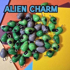 May include: A collection of green and gray alien charm pendants with black eyes. The charms are made of metal and have a shiny finish. The charms are arranged on a yellow, orange, and blue background.