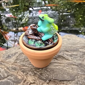 May include: A small terracotta pot topped with a circular lid featuring a sculpted, bright turquoise frog perched on a dark brown textured log. The lid surface is painted with green accents and dark brown details, creating a miniature pond scene.