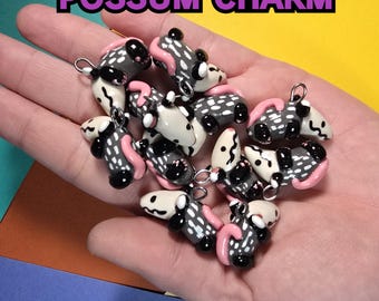 May include: A collection of 10 handmade polymer clay charms shaped like possums. Each charm is painted with black and white spots and has a pink nose and tail. The charms have silver jump rings for attaching to jewelry.