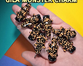 Puede incluir: Una colección de amuletos de monstruo Gila. Cada amuleto es una réplica en miniatura del lagarto, con una base negra y manchas naranjas. Los amuletos se sostienen en una mano, con un fondo amarillo, azul y naranja.