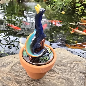 May include: A small, dark blue glass dragon figurine with yellow horns and a curved, multi-colored tail, perched atop a brown stump inside a miniature terracotta pot. The base of the pot contains small green textured elements resembling moss.