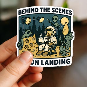 May include: A sticker featuring a cartoon scene with an astronaut, aliens, and a raccoon. The astronaut is seated, holding a mug, with the text "BEHIND THE SCENES ON LANDING" at the top and bottom.