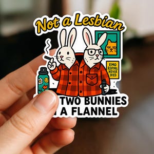 May include: A sticker featuring two cartoon bunnies in a red and black plaid flannel shirt. One bunny smokes a cigarette, and the other wears glasses. Text reads "Not a Lesbian" and "Two Bunnies a Flannel."