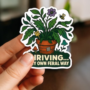 May include: A sticker showing a plant in a terracotta pot with green foliage, a purple flower, and a bee. The pot has a patch. Text reads "THRIVING... MY OWN FERAL WAY."
