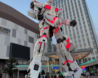 Könnte beinhalten: Eine große, weiße und rote Roboterstatue mit einer langen Antenne steht vor einem modernen Gebäude. Der Roboter hat komplizierte Details und leuchtend rote Akzente. Im Hintergrund sind Menschen versammelt.