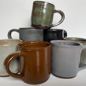 Könnte beinhalten: Eine Sammlung handgefertigter Keramikbecher in verschiedenen Farben, darunter Braun, Grün, Grau und Schwarz. Jeder Becher hat eine einzigartige Form und Glasur, mit einem stabilen Griff. Die Becher sind gestapelt und vor weißem Hintergrund angeordnet.
