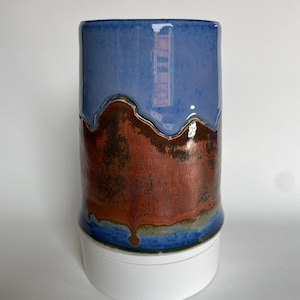 May include: A handmade ceramic tumbler with a glossy finish. The top half is a solid blue, transitioning to a textured, reddish-brown middle section. The base features a band of blue and green. The tumbler sits on a white base.