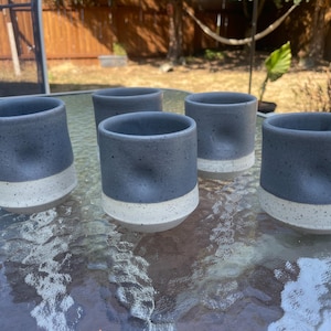 May include: A set of five ceramic cups featuring a two-tone design with a dark gray speckled upper section and a solid off-white base. Each cup has a distinct thumb-sized indentation on the side, providing a textured grip for the user.