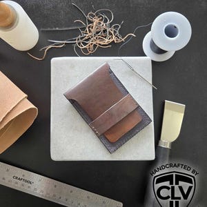 May include: A brown leather wallet with a black leather trim. The wallet is handmade and features the logo "CLV Clay Legacy Ventures".