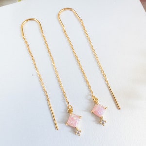 May include: A pair of gold chain earrings with pink square-shaped stones and a small clear stone accent. The earrings are dangling and have a delicate chain design.