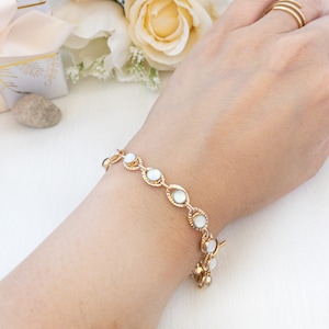 May include: Gold-toned bracelet featuring oval, iridescent stones. The bracelet is connected by a delicate chain and has a small charm at the end. The bracelet is worn on a wrist, with a background of flowers and a gift box.