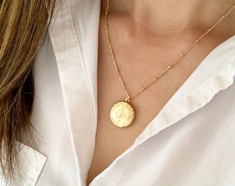 May include: A gold-colored necklace with a round pendant featuring a detailed design. The necklace is worn over a white collared shirt. The chain has small, evenly spaced beads. The pendant has a detailed relief design.