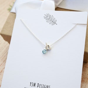 May include: A silver necklace with a cursive letter 'b' charm and a light blue gemstone pendant. The necklace is displayed on a white card with the text "YSM DESIGNS HANDCRAFTED WITH LOVE".