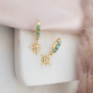 May include: A pair of gold-colored hoop earrings with turquoise-colored beads and a starburst charm. The earrings are displayed on a white surface with a soft pink background and a feathery plant.