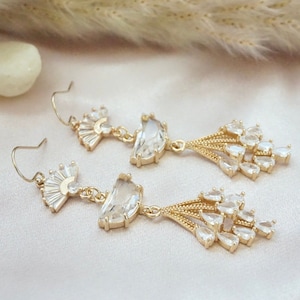 May include: A pair of gold tone dangle earrings with clear crystal accents. The earrings feature a fan-shaped design with a larger crystal in the center.