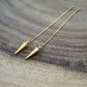 May include: A pair of gold dangle earrings with a pointed, cone-shaped design. The earrings are suspended from a delicate gold chain.