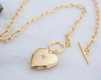 May include: Gold-colored heart-shaped locket necklace with a small diamond-like accent. The locket is on a gold chain with a toggle clasp. The necklace is displayed on a white marble surface.