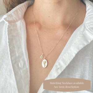 May include: A gold chain necklace with a white oval pendant featuring a yellow flower design. The pendant also has a small gold letter "B" charm attached to it.