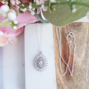 May include: A silver necklace with a pendant featuring a central marquise-cut stone surrounded by smaller, round, clear stones. The pendant is on a delicate silver chain. The necklace is displayed on a white surface with pink and green floral accents.
