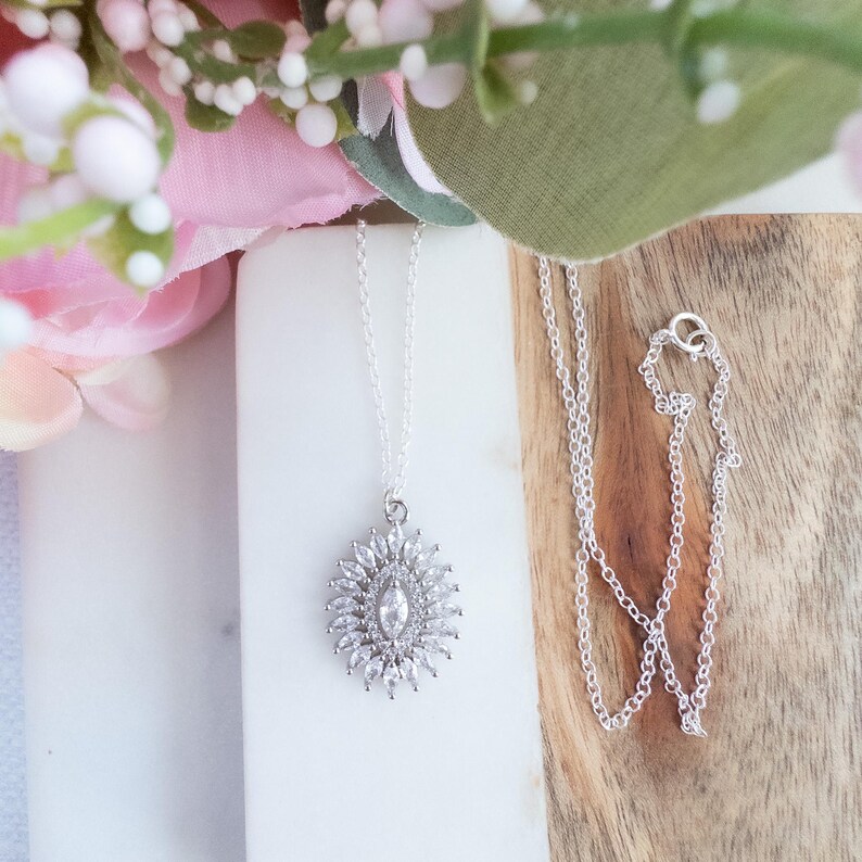 May include: A silver necklace with a pendant featuring a central marquise-cut stone surrounded by smaller, round, clear stones. The pendant is on a delicate silver chain. The necklace is displayed on a white surface with pink and green floral accents.