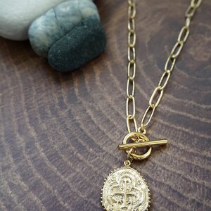 May include: A gold chain necklace with a toggle clasp and a large oval pendant featuring a cross design.
