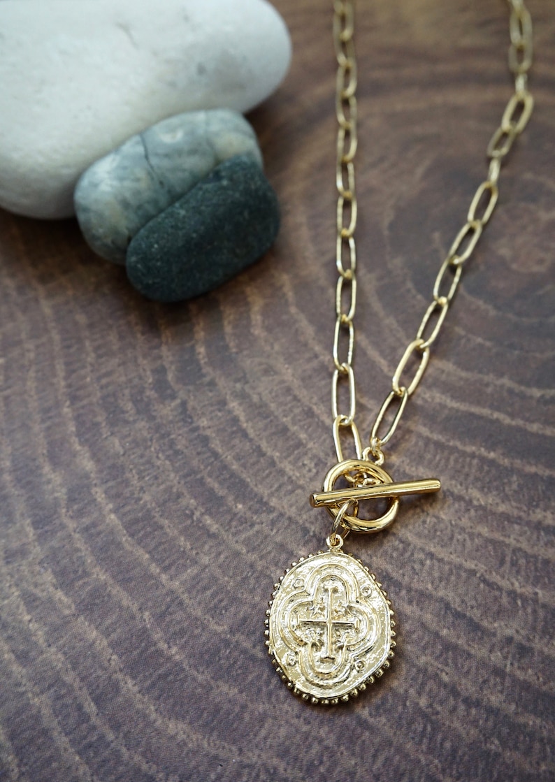 May include: A gold chain necklace with a toggle clasp and a large oval pendant featuring a cross design.