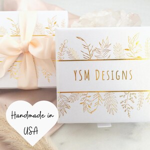 May include: White gift boxes with gold leaf designs and the text "YSM DESIGNS". One box is tied with a peach ribbon. A heart-shaped sticker reads "Handmade in USA".